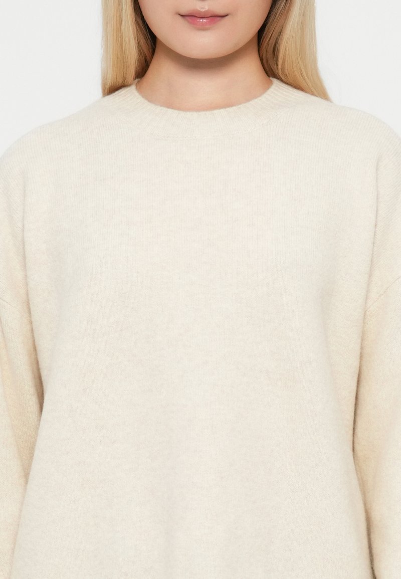 Sweter w kolorze kremowym o miękkiej teksturze, okrągłym dekolcie i opadających ramionach, z prostym, minimalistycznym wzorem.