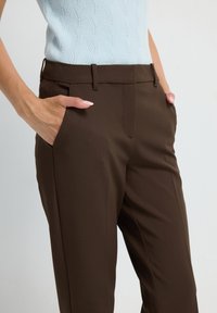 Pantalons marron ajustés avec une texture lisse, dotés de poches latérales et d'une fermeture éclair à l'avant, associés à un haut tricoté bleu clair.