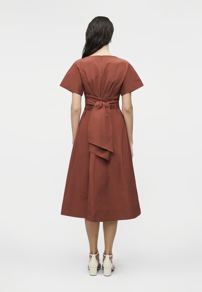 Γυναίκα με μακριά σκούρα μαλλιά, που φοράει μεσομήκη φόρεμα σε σκουριά απόχρωση με κοντά μανίκια, φερμουάρ στην πλάτη και λεπτομέρεια δέσιμου υφάσματος στη μέση, λευκές γόβες.