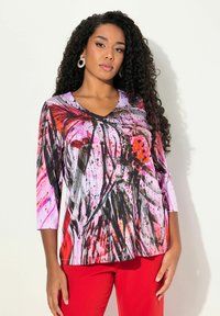 Langærmet V-hals top i farverigt abstrakt print med livlige pink, sorte og røde sprøjt på et blødt stof, parret med røde bukser.