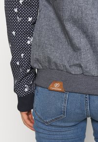 Personne portant un jean bleu et une veste grise et bleu marine avec des motifs floraux sur la manche et une étiquette marron "ragwear" sur la ceinture.
