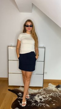 Bela majica s kratkimi rokavi, mornarsko modra krila s čepi, bele sandale s peto in črne sončna očala, stoji na nevtralni podlagi.