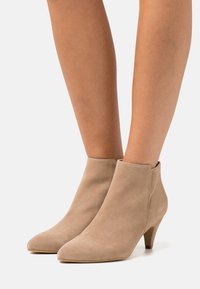Beige ruskind ankelstøvler med spids tå og lav hæl. Glat tekstur og minimalistisk design med en buet silhuet.