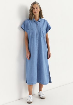 Vrouw met schouderlang grijs haar draagt een lichtblauwe midi spijkerjurk met knopen en witte sneakers, staand tegen een witte muur.