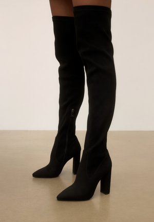 Bottes noires en daim au-dessus du genou à talons hauts avec fermeture éclair latérale portées sur des jambes nues sur un fond uni.