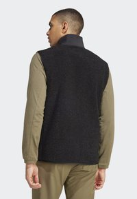 Schwarze Fleece-Weste mit hohem Kragen, aus strukturiertem Stoff und glattem, kontrastierendem Vorderteil, kombiniert mit einem olivgrünen Langarmshirt.