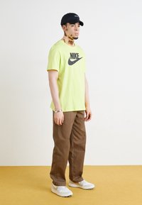 T-shirt Nike verde néon com um logo preto, calças castanhas e ténis brancos. A pessoa usa um chapéu preto e uma pulseira prateada.