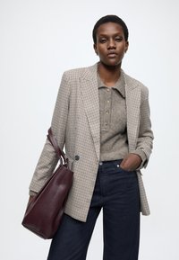 Kariran blazer v bež in rjavi barvi, siva pletena polo majica, temne kavbojke iz jeansa in bordo usnjena torbica z teksturirano površino.