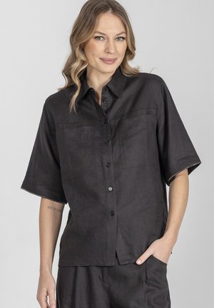 Kobieta o blond falowanych włosach nosi luźną czarną koszulę na guziki z krótkim rękawem i pasujące spodnie, pozując z jedną ręką w kieszeni.