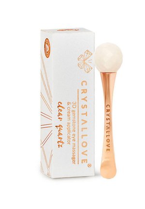 Un masseur pour les yeux en quartz clair avec une poignée en or rose, accompagné d'une boîte blanche ornée de motifs géométriques et de texte pour l'identification du produit.