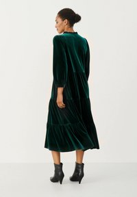 Groene fluwelen jurk met lange mouwen en een gelaagde rok. De jurk heeft een ronde halslijn en wordt gecombineerd met zwarte enkellaarzen.
