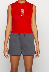 Red sleeveless crop top with a ribbed texture and "TOMMY" logo centered. Paired with gray, drawstring elastic shorts.