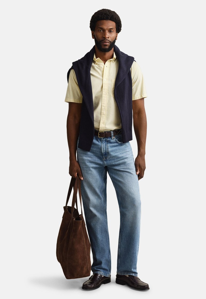 Hombre con camisa amarilla de manga corta y un suéter azul marino sobre los hombros, vaqueros azules, zapatos oscuros, sosteniendo un gran bolso marrón.