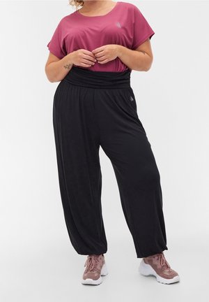 Vrouw draagt losse zwarte broek, een maroonkleurige korte mouw top en roze sneakers, staat met de handen samengevouwen ter hoogte van de taille.