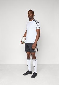 Hvitt treningstrøye med svarte striper, svarte shorts med gullakenter, svarte fotballsko og hvite sokker med svarte striper. Holder en fotball.