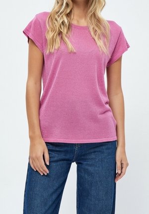 Kobieta ubrana w różowy, teksturowany t-shirt z krótkim rękawem i niebieskie jeansy denim, stojąca w luźnej pozie na jednolitym tle.