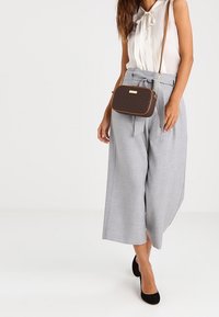 Donna che indossa una blusa bianca senza maniche, pantaloni grigi a vita alta e larghi con cintura, tacchi neri e una borsa a tracolla marrone di design.