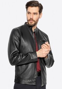 Giubbotto in pelle nera con collo a listino e chiusura con zip, dotato di taschino sul petto e texture liscia. Design slim fit, stile minimalista.