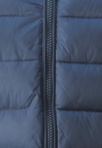 Primo piano di giacca imbottita marina, con pannelli trapuntati, cerniera centrale con denti in plastica nera e tessuto liscio e strutturato.