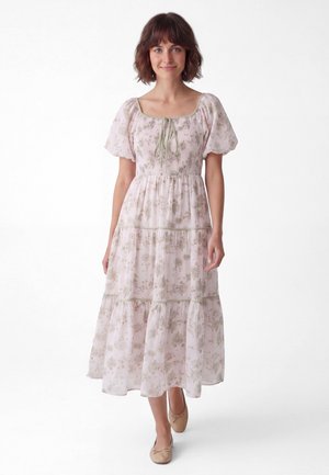 Jonge vrouw met kort bruin haar, die een lichte bloemenjurk met pofmouwen en beige balletflatjes draagt, staand tegen een witte achtergrond.