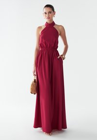 Langes Maxikleid mit Halterneck aus tiefrotem Stoff, mit elastischem Bund und seitlichen Taschen, kombiniert mit einer kleinen braunen Handtasche.