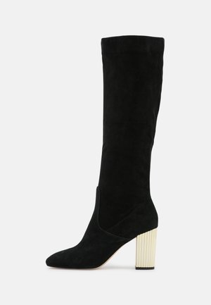 Bottine haute en daim noir jusqu'au genou avec bout pointu et talon bloc côtelé de couleur crème, inclinée vers la droite sur un fond blanc.