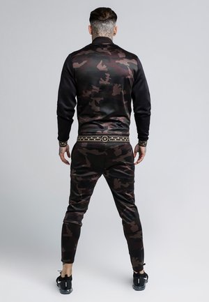 Mężczyzna stojący tyłem, ubrany w czarną i ciemnobrązową kurtkę i spodnie w wzór moro, ze złotym wzorem na pasku w talii oraz czarne trampki.