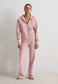 Femme debout pieds nus contre un mur blanc, portant un hoodie rose à fermeture éclair et un pantalon large assorti.