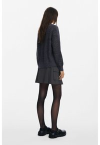 Pull en maille gris foncé à manches longues, associé à une jupe plissée noire avec poches latérales, des collants opaques et des chaussures épaisses.