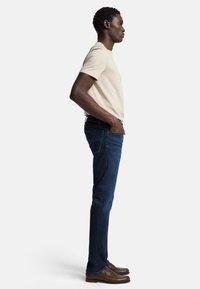 Beige T-Shirt, figurbetont, kombiniert mit dunkelblauen Jeans. Das Model steht seitlich, die Hände in den Taschen, trägt braune Schuhe mit dezenter Verzierungen.