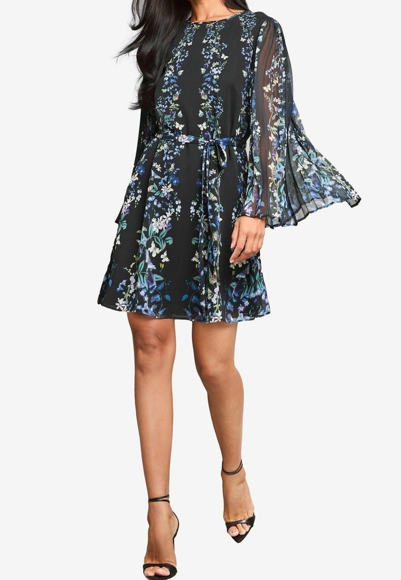 Femme portant une robe noire à fleurs avec des manches longues plissées transparentes, une ceinture nouée à la taille et des sandales noires à talons hauts avec bride à la cheville.