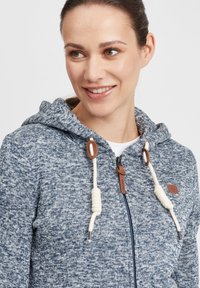 Mikina s kapucňou na zips z modrej pleteniny so zmysluplnou textúrou, s krémovými šnúrkami, koženými doplnkami a detailom loga.
