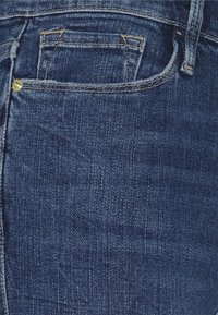Jeans de mezclilla azul oscuro con costuras visibles, un pequeño bolsillo en la parte delantera y un detalle de botón en el lado derecho. Textura suave.