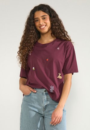 Jeune femme aux cheveux bouclés portant un t-shirt bordeaux avec de petits patchs sur le thème de la nature et un jean bleu clair. Elle sourit, une main dans la poche.