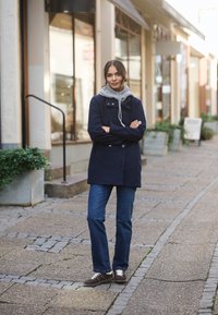 Marineblauer Doppelreiher-Mantel mit grauem Kapuzenpullover, blauen Jeans und braunen Sneakers; mit Knöpfen und Vordertaschen, auf einer Kopfsteinpflasterstraße.