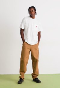 White short-sleeve t-shirt with a chest pocket and red logo, paired with tan loose-fitting trousers and black shoes, set against a neutral background.