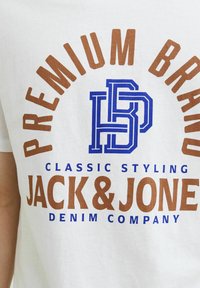 Biele bavlnené tričko so výrazným hnedým a modrým textom. Dizajn obsahuje nápis "PREMIUM BRAND" a "JACK & JONES DENIM COMPANY" s grafickými logovými akcentmi.