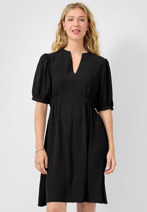 Vrouw met golvend blond haar, gekleed in een zwarte jurk tot op de knie met pofmouwen, een gerimpelde taille en een gespleten halslijn, staand tegen een effen achtergrond.