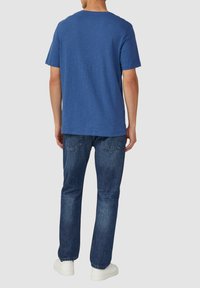 Blauwe T-shirt met korte mouwen en een relaxte pasvorm, gecombineerd met donkere spijkerbroeken en witte sneakers, bekeken vanuit de achterkant.