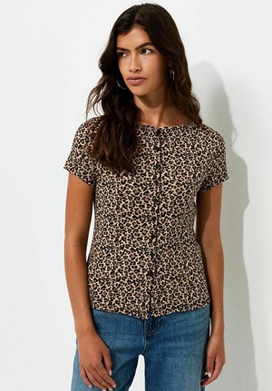Vrouw met lang donker haar draagt een kort mouwtjes blouse met luipaardprint en blauwe spijkerbroek, kijkt iets naar de zijkant tegen een witte achtergrond.