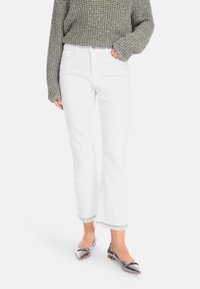 Weiße Schlagjeans mit fransigem Saum, kombiniert mit einem locker gestrickten grauen Sweater. Die Schuhe sind glänzende, spitze Ballerinas mit metallischem Finish.