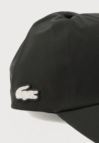 Melna cepure no gludas auduma ar apaļu augšu. Sānā ir rhinestone logotipa aplikācija. Vizīre ir taisna un teksturēta.