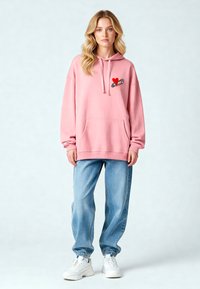 Felpa con cappuccio rosa presenta un piccolo design di un cuore rosso e una spilla di sicurezza sul petto; abbinata a jeans a gamba stretta blu chiaro e sneakers bianche.