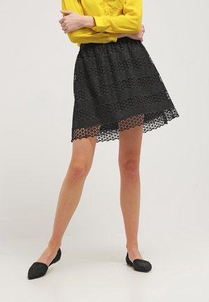 Musta pitsihame, jossa on kerroksittainen muotoilu ja helmassa leikatut geometriset kuviot. Yhdistetty keltaiseen puseroon ja mustiin tasapohjaisiin kenkiin.