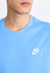 Nebesno modra bombažna majica s krožnim izrezom, ki ima izvezen bel Nike logotip na levi strani prsnega dela. Gladka tekstura.
