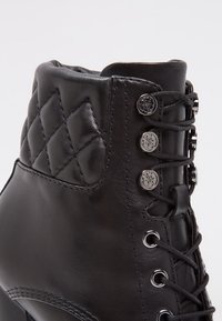 Bottine en cuir noir avec texture matelassée, garniture argentée et design à lacets. Elle présente un bout arrondi et une finition lisse.