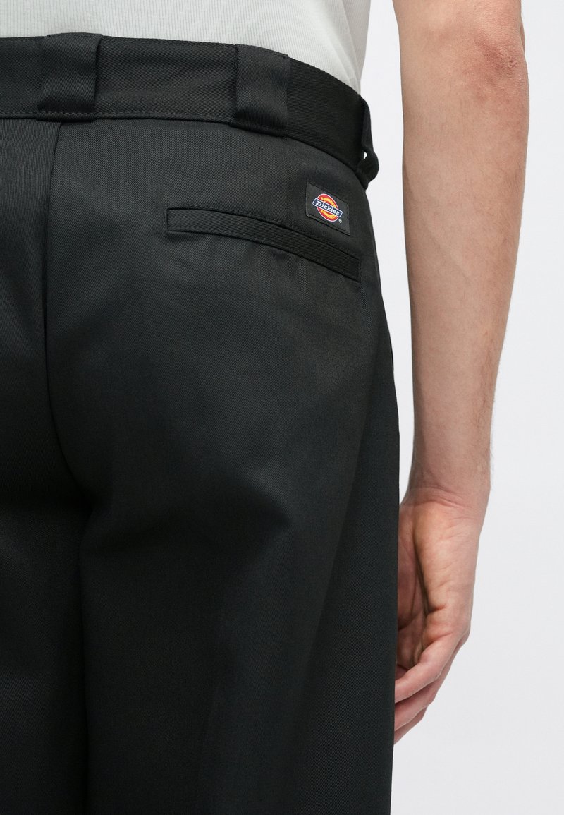 Sorte bukser med en glat tekstur, der har en baglomme og et mærke i rød, blå og gul på taljen.