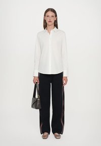 Camicia bianca con bottoni, pantaloni neri a gamba larga con strisce laterali rosse e bianche, e sandali con motivo; porta una borsa grigia.