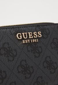 Pochette noire texturée avec le logo en or "GUESS EST 1981". Présente un motif subtil et une fermeture éclair sur le bord supérieur. Matériel durable.