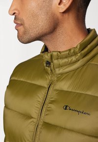 Olivgrüne Steppjacke mit Stehkragen, vertikaler Quilt-Design, schwarzem Reißverschluss und gesticktem "Champion"-Logo auf der Brust.